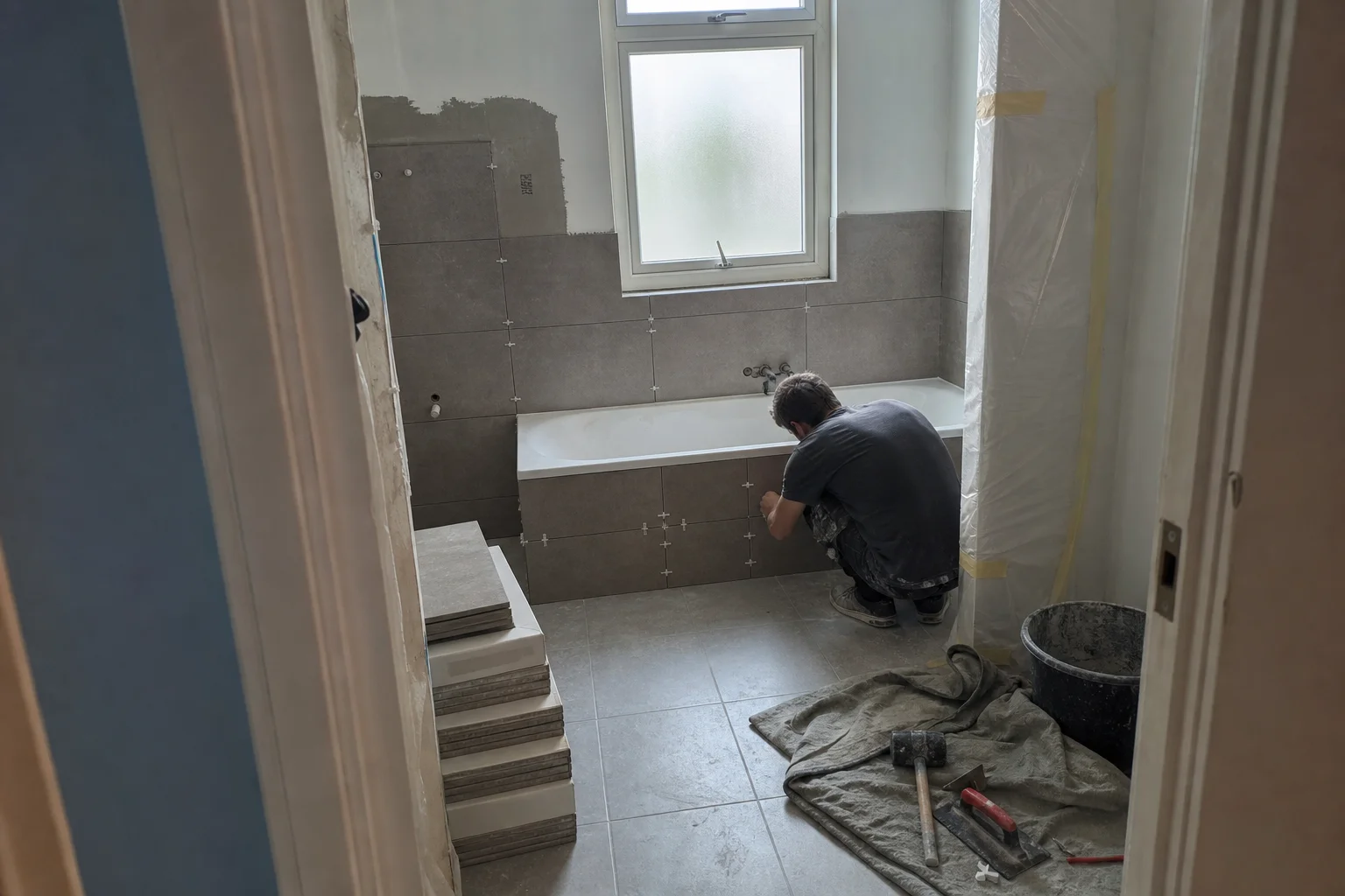 Halvfärdig badrumsrenovering utan bygglov, med kaklade väggar, verktyg och en person som arbetar vid badkaret.